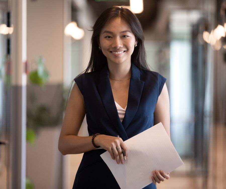 Woman in office corridor smiling while carrying some paperwork.