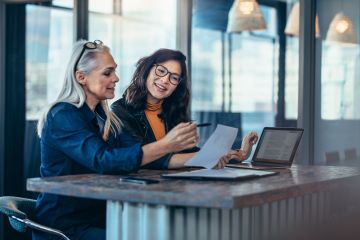 Two females in corporate environment reviewing printed and digital documents. 