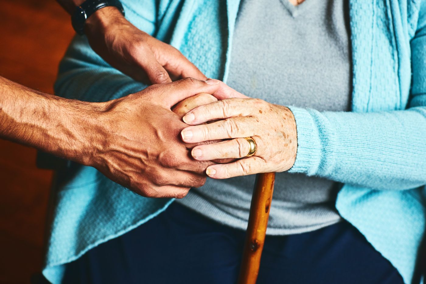 Young male hands gently cupping hands of frail elderly seated lady.