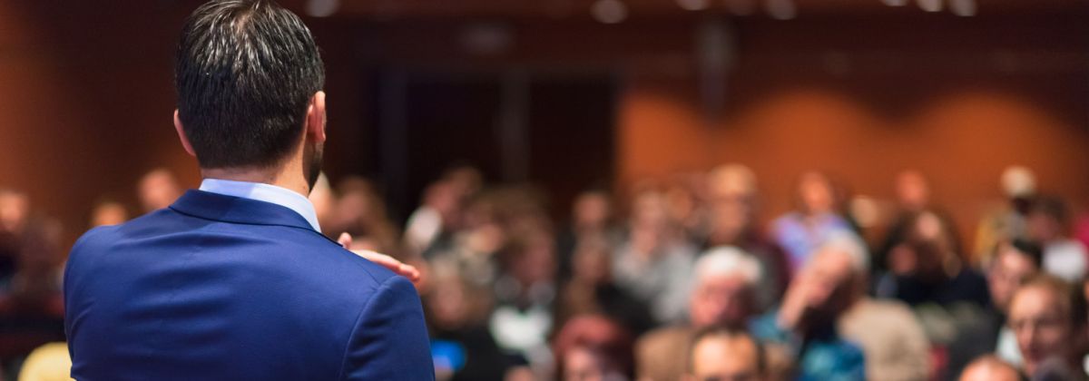 Man in a blue suit giving a presentation to a large, blurred audience in a conference room.